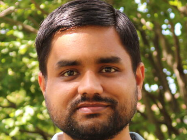 Kaustubh is in front of bushes and greenery and smiles at the camera